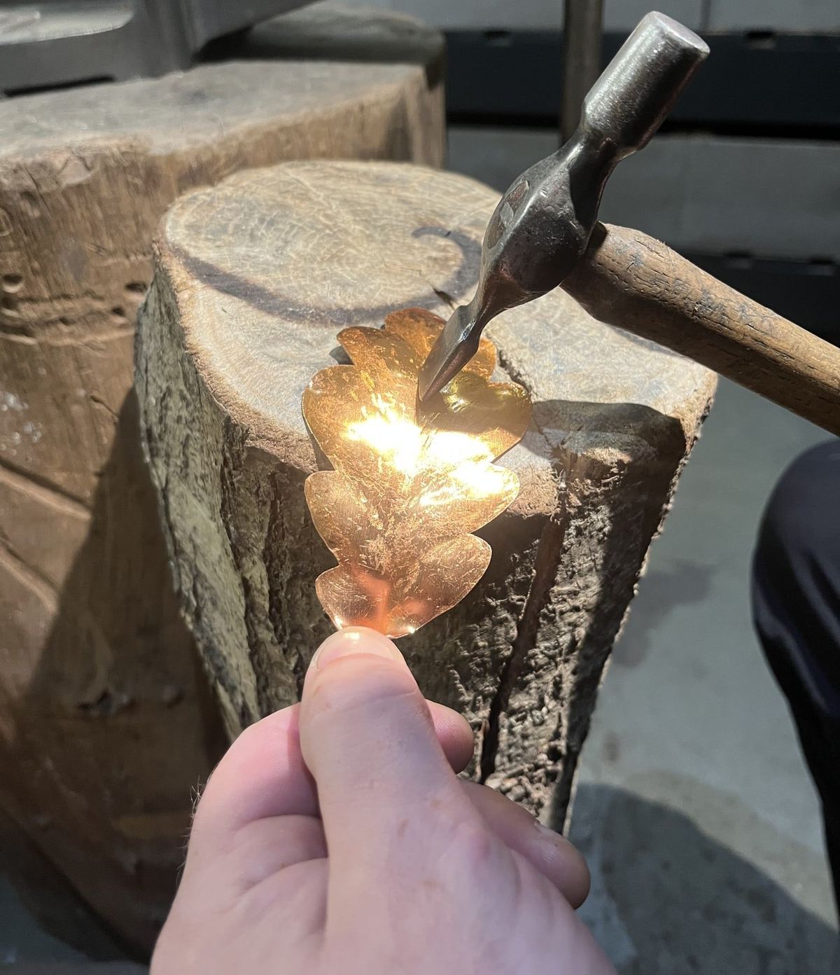 A hand holding a copper oak leaf on the stump while the hammer shapes it, the metal glowing